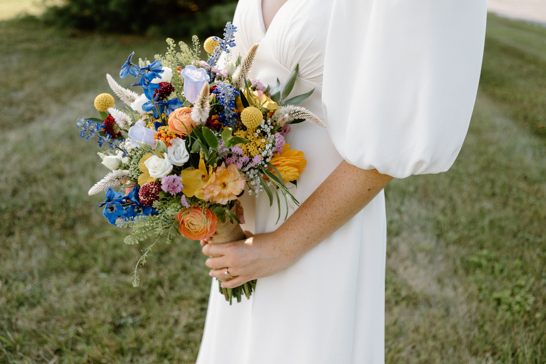 Bride outdoors on grass holding vivid bouquet of blue delphinium, orange ranunculus, yellow billy buttons