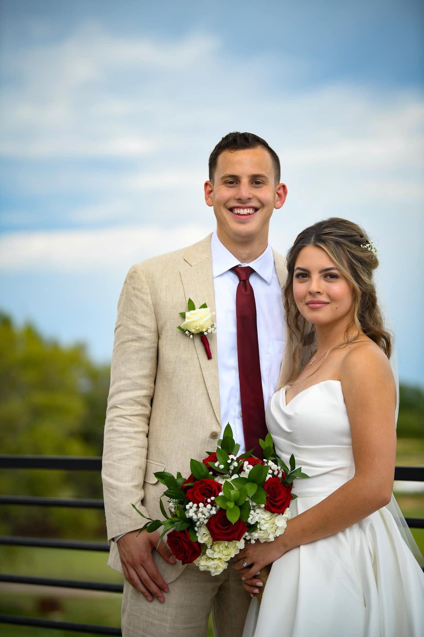 Bride holding deep red rose bouquet with baby's breath, beside groom wearing white rose boutonniere