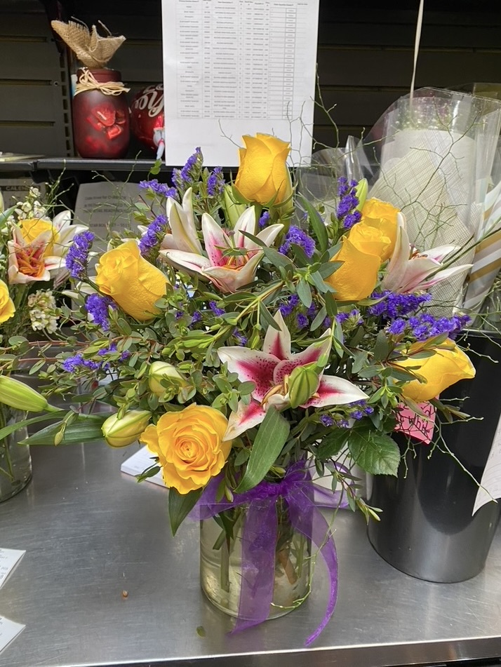 Mason jar arrangement of yellow roses, pink lilies and purple statice on worktable