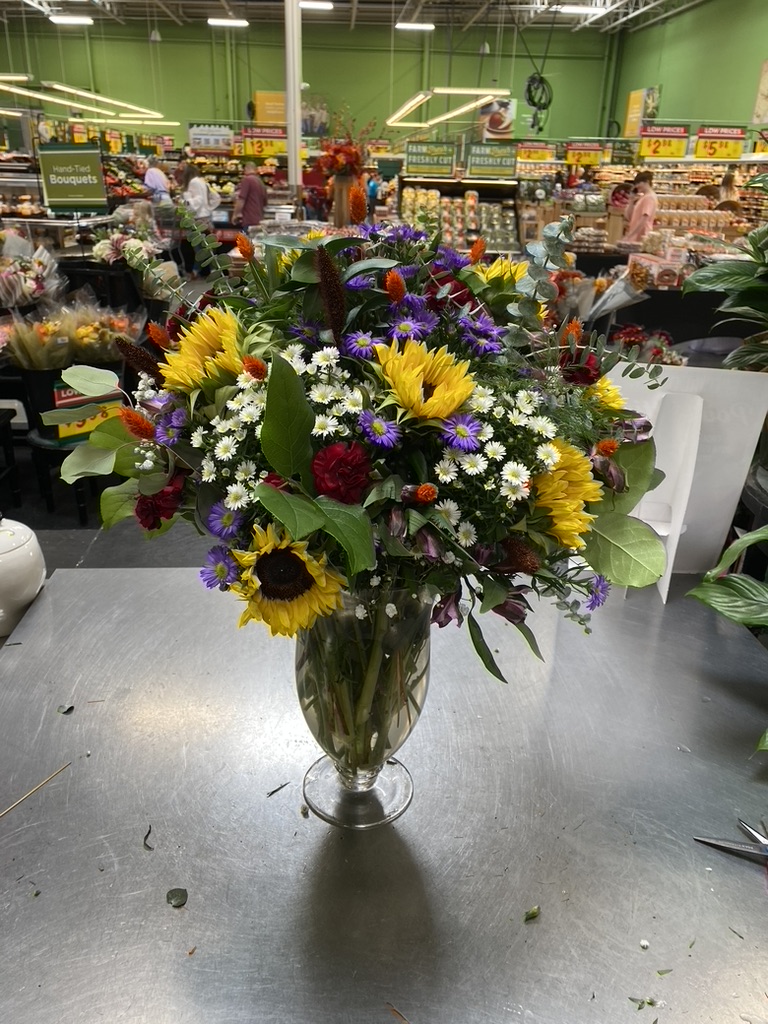 Sunflowers, purple asters and white daisies in footed glass vase on florist counter