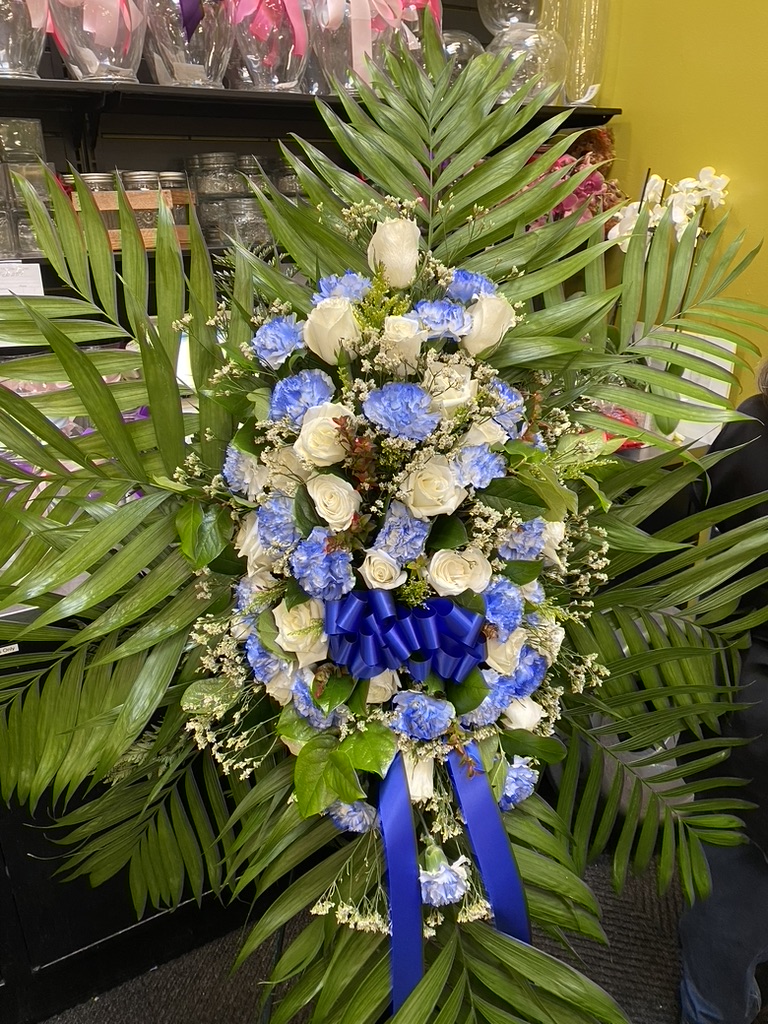Standing sympathy spray of blue carnations, white roses, palm fronds, blue ribbon