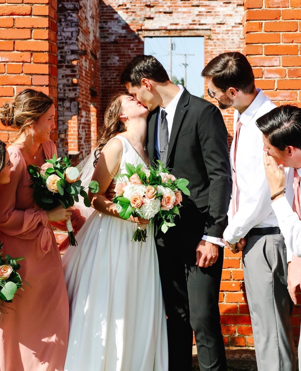 Couple kissing outside brick building, bride carrying blush and peach rose bouquet with eucalyptus
